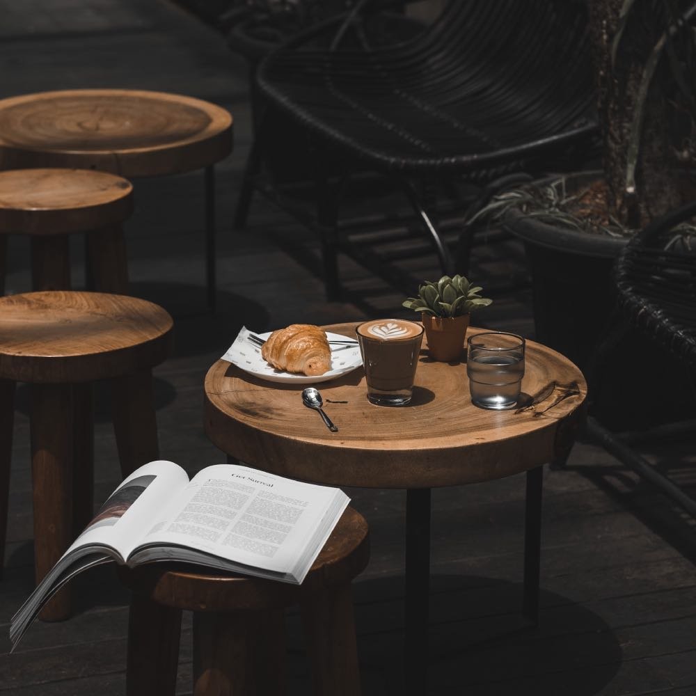 Wooden table featuring a cup of coffee, a croissant, a glass of water, and an open book.