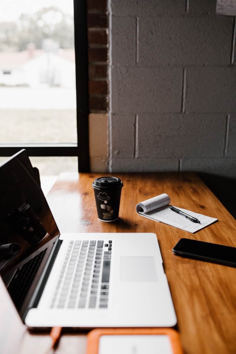 A laptop, coffee cup, notepad, and pen on a wooden table in a casual setting.